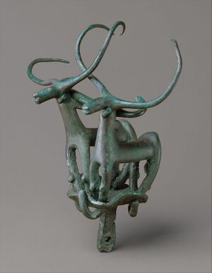 An image of a Copper-alloy finial depicting a pair of long-horned bulls, likely part of a ceremonial standard. The exaggerated curving horns and stylized forms reflect Mvanagazi highland artistic conventions and their association with fertility and ritual power.